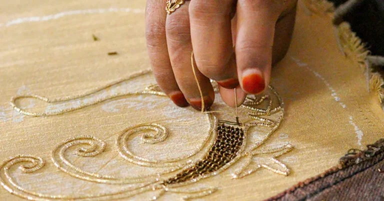 Artisan's hands working gold zardozi metal-thread embroidery on fabric, couching coiled dabka wire
